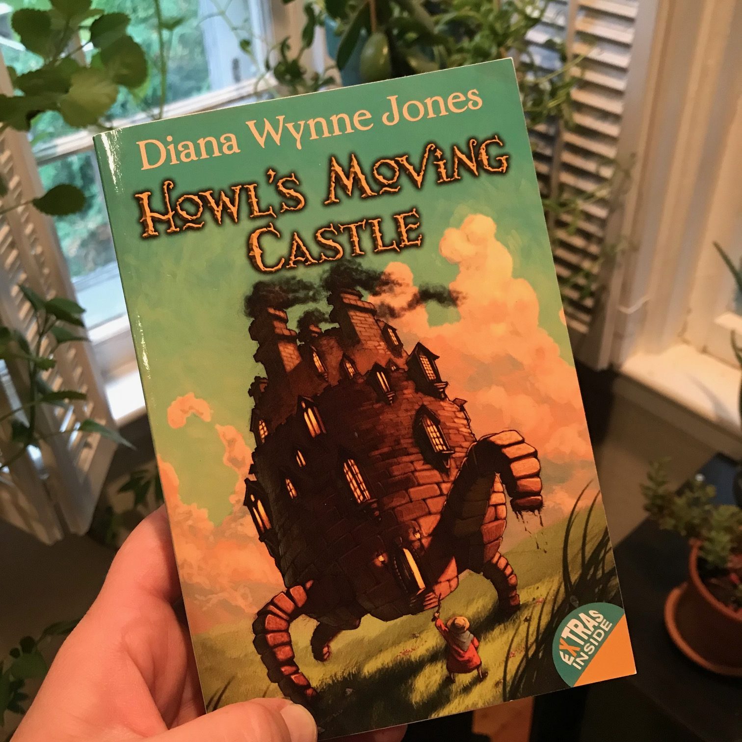 Cover of the book "Howl's Moving Castle" with a brick castle with four legs walking across the grass, and a figure seen from behind holding up a stick toward it.