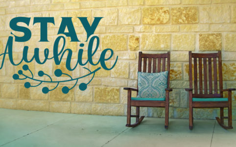 Two rocking chairs in front of a brick wall with the text "stay awhile" to the left