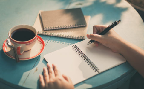 open notebook with hands holding a pen and on a teal table with a cup of tea and two other notebooks stacked nearby editing for self publishing